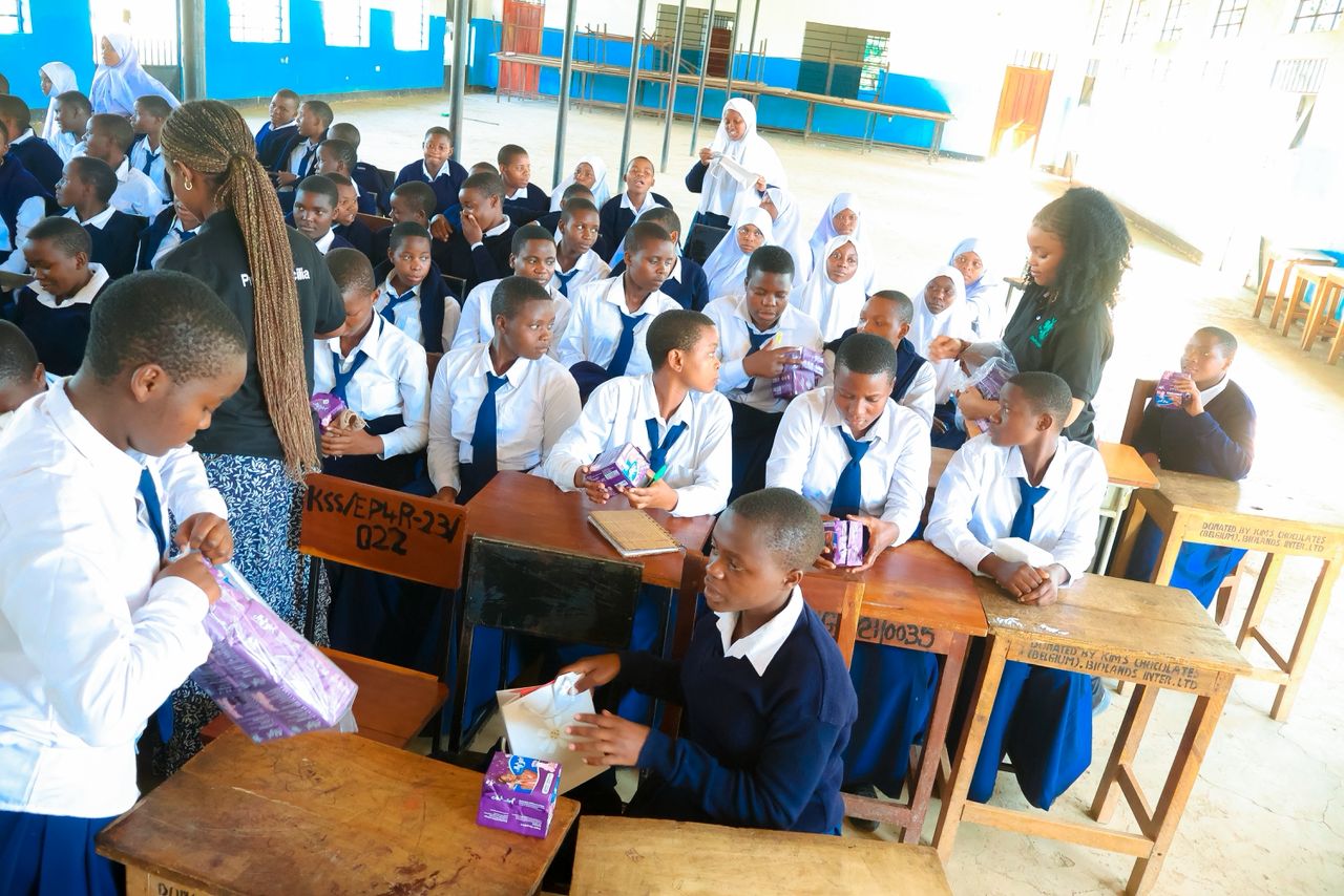 Students at their desks holding sanitary pad packets as they are handed out in the school hall