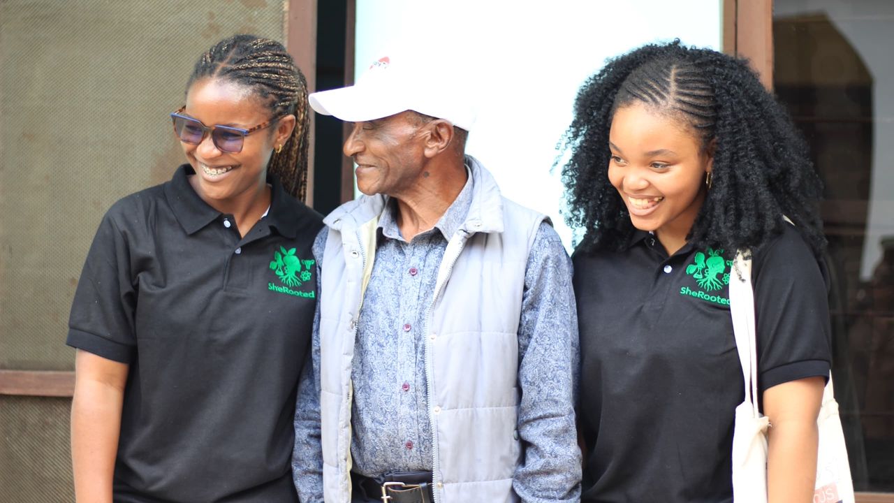 Two SheRooted volunteers smiling beside an older man at Kafundo Secondary School